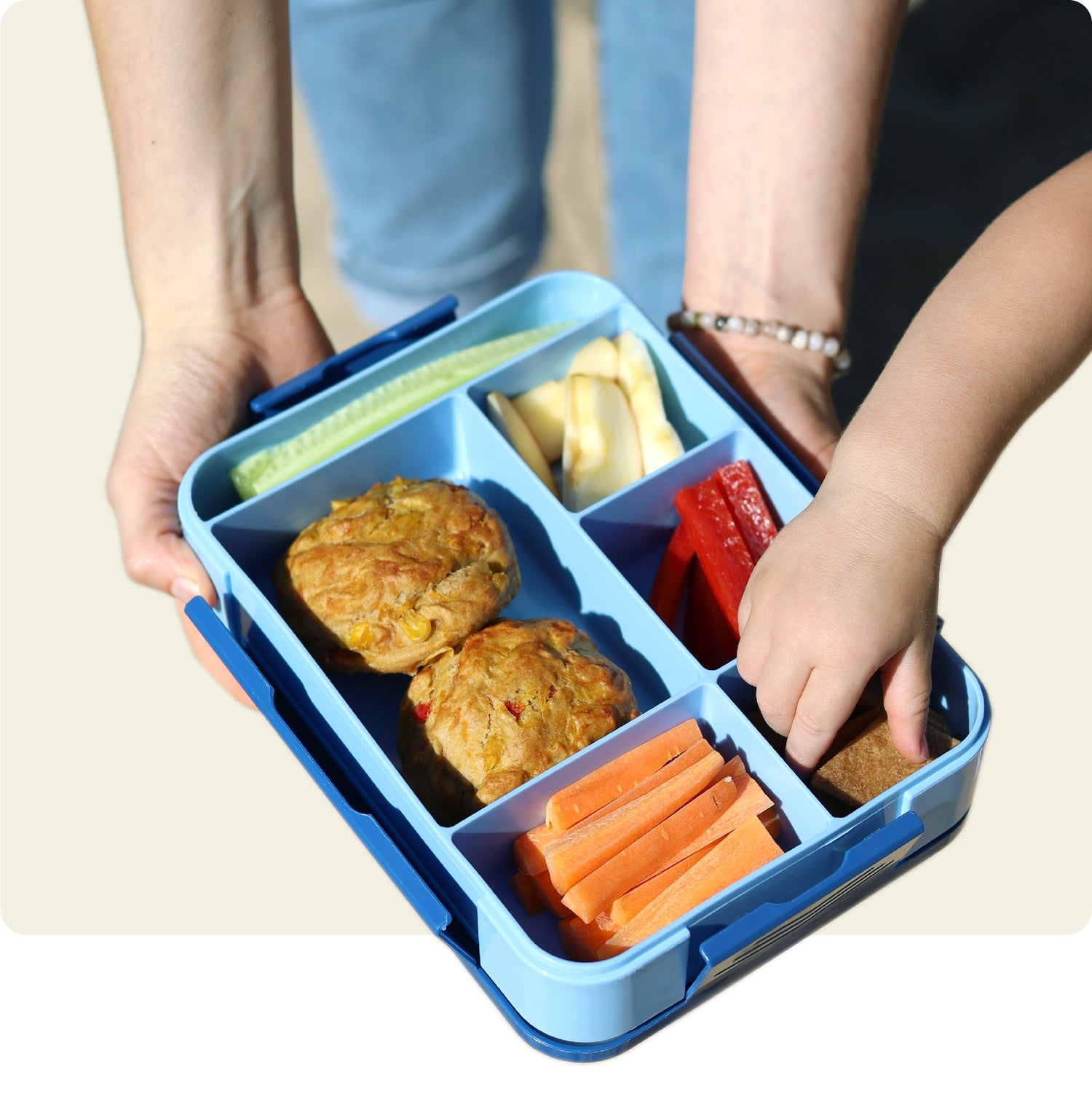 Mutter bietet Kind gesunde, vielfältig gefüllte Brotdose mit Snacks an, Kind bedient sich mit Freude.