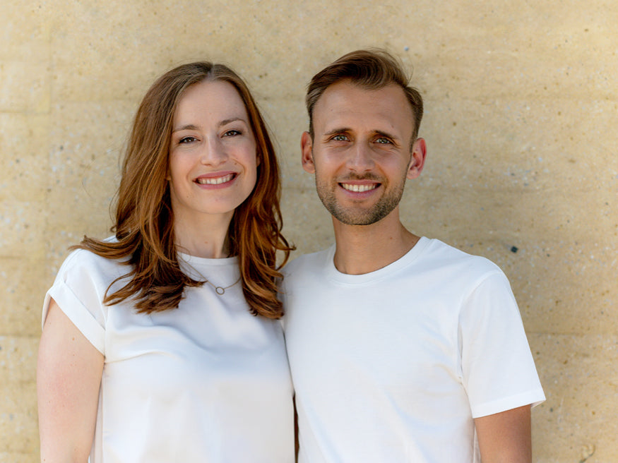 Clemens und Sophie, Mediziner und Experten für gesunde Kinderernährung, lächeln vor sandfarbenem Hintergrund.